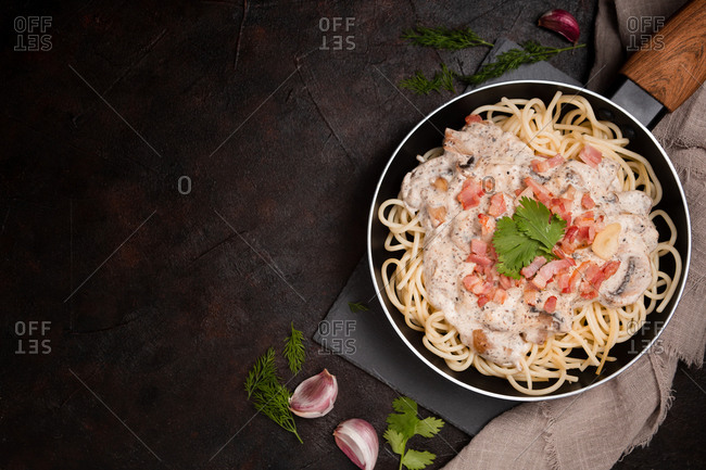 Composition of tasty spaghetti with ham slices and mushrooms in creamy sauce cooked in pan and placed on wooden cutting board at dark table with garlic and gray linen fabric aside