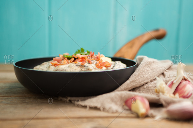 Composition of tasty spaghetti with ham slices and mushrooms in creamy sauce cooked in pan and placed on wooden cutting board at a wooden table with garlic and linen fabric aside on blue background