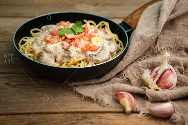 From above tasty spaghetti with ham slices and mushrooms in creamy sauce cooked in pan and placed on wooden cutting board at a wooden table with garlic and linen fabric aside