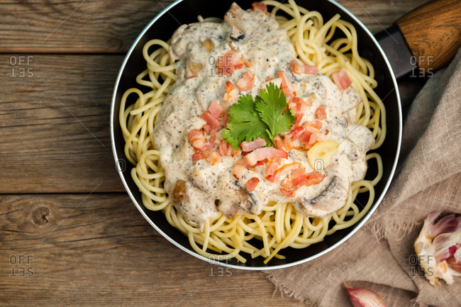 From above tasty spaghetti with ham slices and mushrooms in creamy sauce cooked in pan and placed on wooden cutting board at a wooden table with garlic and linen fabric aside