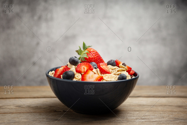 From above delicious breakfast bowl of corn flakes with strawberries and blueberries placed on cutting board and decorated with linen cloth and berries around dish on wooden table with gray background
