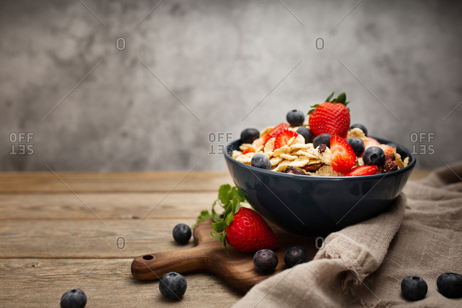 From above delicious breakfast bowl of corn flakes with strawberries and blueberries placed on cutting board and decorated with linen cloth and berries around dish on wooden table with gray background