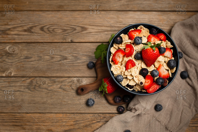 Top view of delicious breakfast bowl of corn flakes with strawberries and blueberries placed on cutting board and decorated with linen cloth and berries around dish on wooden background