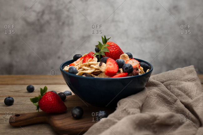 From above delicious breakfast bowl of corn flakes with strawberries and blueberries placed on cutting board and decorated with linen cloth and berries around dish on wooden table with gray background