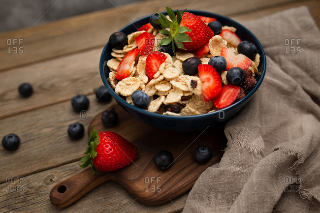 From above delicious breakfast bowl of corn flakes with strawberries and blueberries placed on cutting board and decorated with linen cloth and berries around dish on wooden background