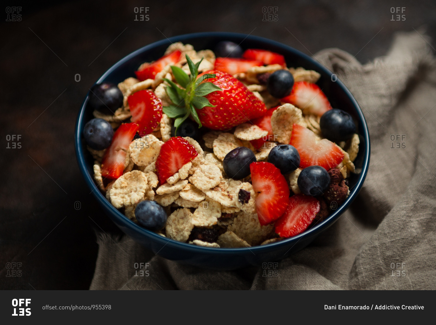 Top view of delicious breakfast bowl of corn flakes with strawberries and blueberries placed on cutting board and decorated with linen cloth and berries around dish on wooden background