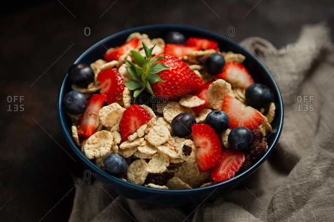 Top view of delicious breakfast bowl of corn flakes with strawberries and blueberries placed on cutting board and decorated with linen cloth and berries around dish on wooden background