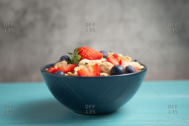 Delicious breakfast bowl of corn flakes with strawberries and blueberries placed on cutting board and decorated with linen cloth and berries around dish on blue wooden table with gray background