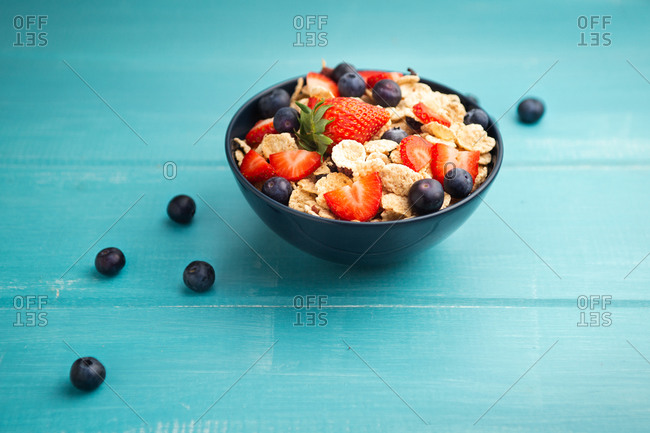 From above delicious breakfast bowl of corn flakes with strawberries and blueberries placed on cutting board and decorated with linen cloth and berries around dish on wooden blue background