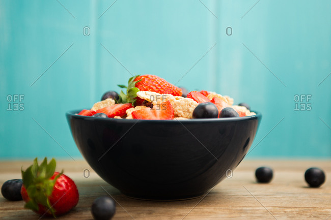 From above delicious breakfast bowl of corn flakes with strawberries and blueberries placed on cutting board and decorated with linen cloth and berries around dish on wooden table with blue background