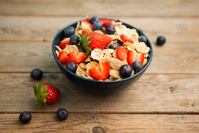 Top view of delicious breakfast bowl of corn flakes with strawberries and blueberries placed on cutting board and decorated with linen cloth and berries around dish on wooden background