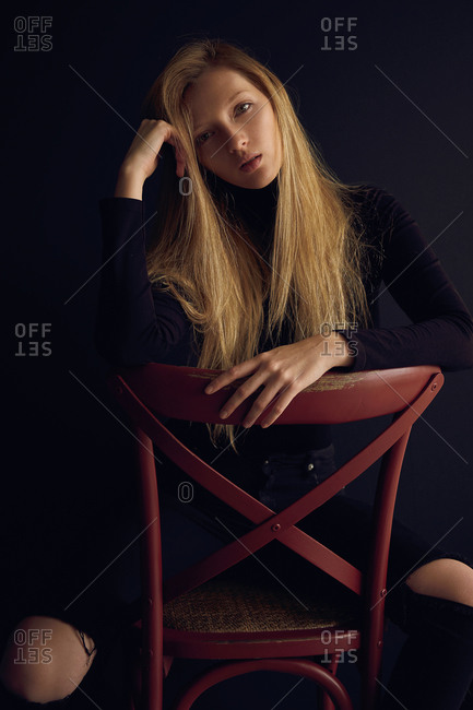 Young blond woman in dark clothes sitting on chair against black background and looking away