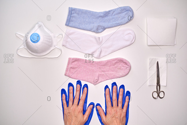Top view of unrecognizable person hands on top of blue gloves near sock, smartphone, napkin, mask and scissors to make fabric mask during quarantine period of coronavirus for protection