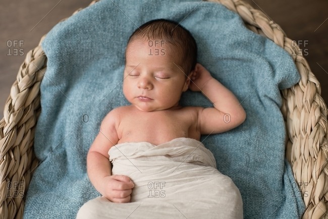 Top view of newborn baby wrapped in cloth lying on soft blanket and sleeping in wicker basket on floor at home
