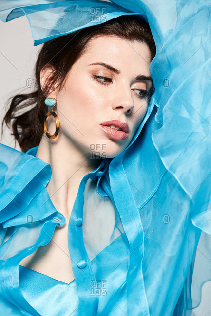 Alluring model wearing transparent blue blouse and covering face with textile while looking at camera in studio