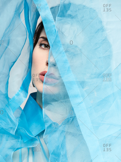 Alluring model wearing transparent blue blouse and covering face with textile while looking at camera in studio