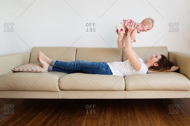 Smiling mother lying on couch lifting her baby above her