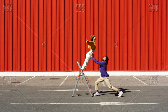 Young man and woman performing with a ladder in front of a red wall