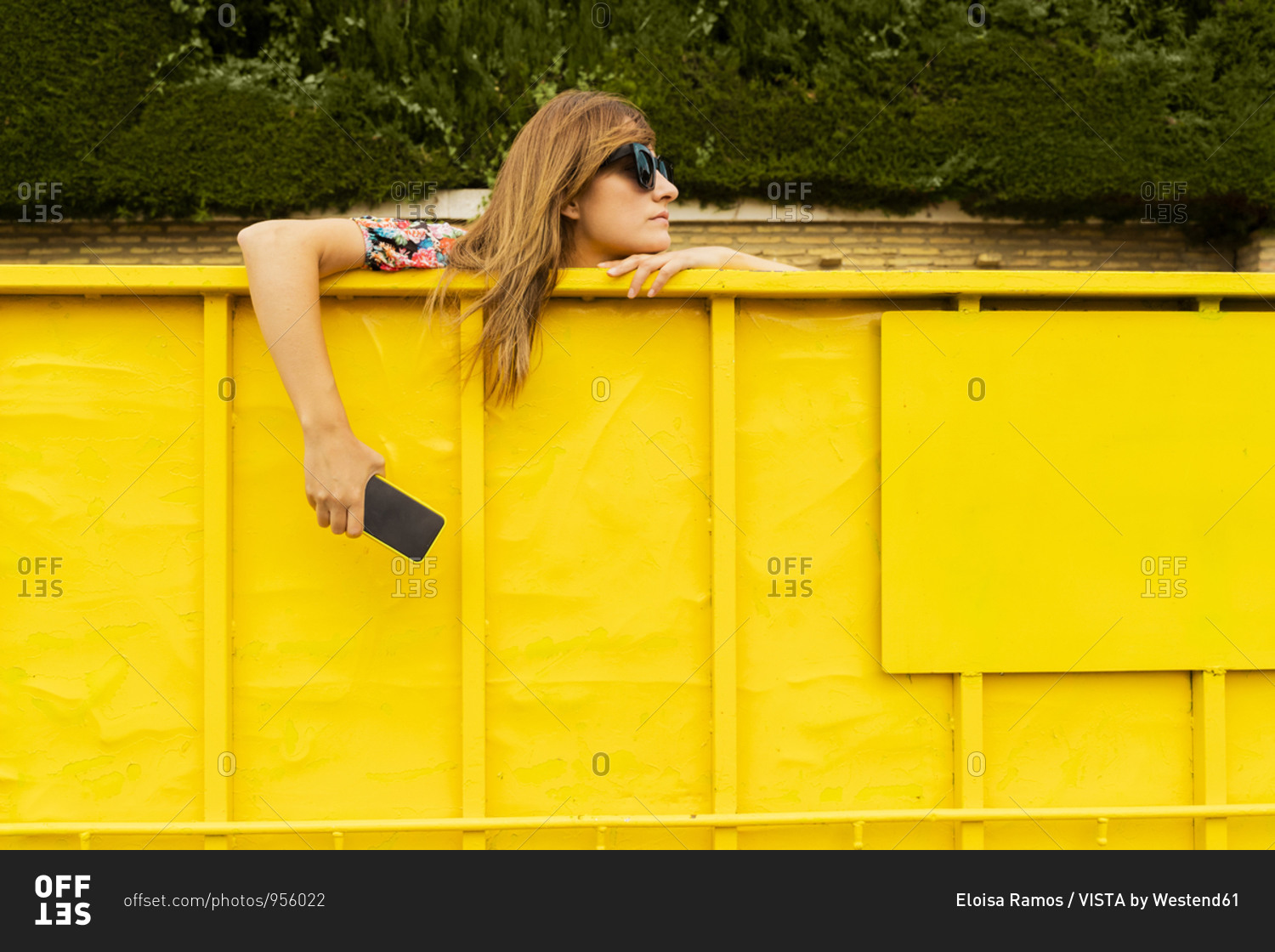 Woman wearing sunglasses- leaning on edge on yellow container- holding smartphone