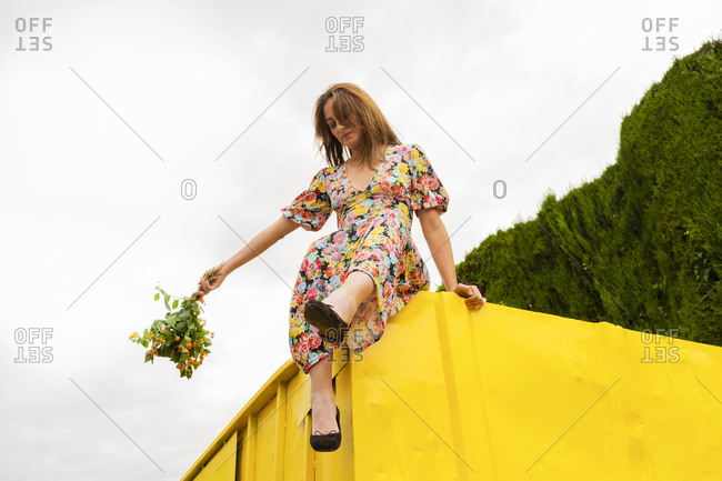 Woman in flower dress sitting on edge of yellow container- holding bunch of flowers