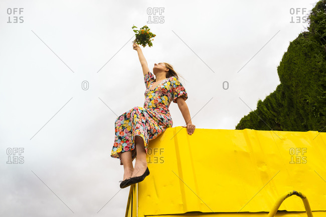 Woman in flower dress sitting on edge of yellow container- holding bunch of flowers