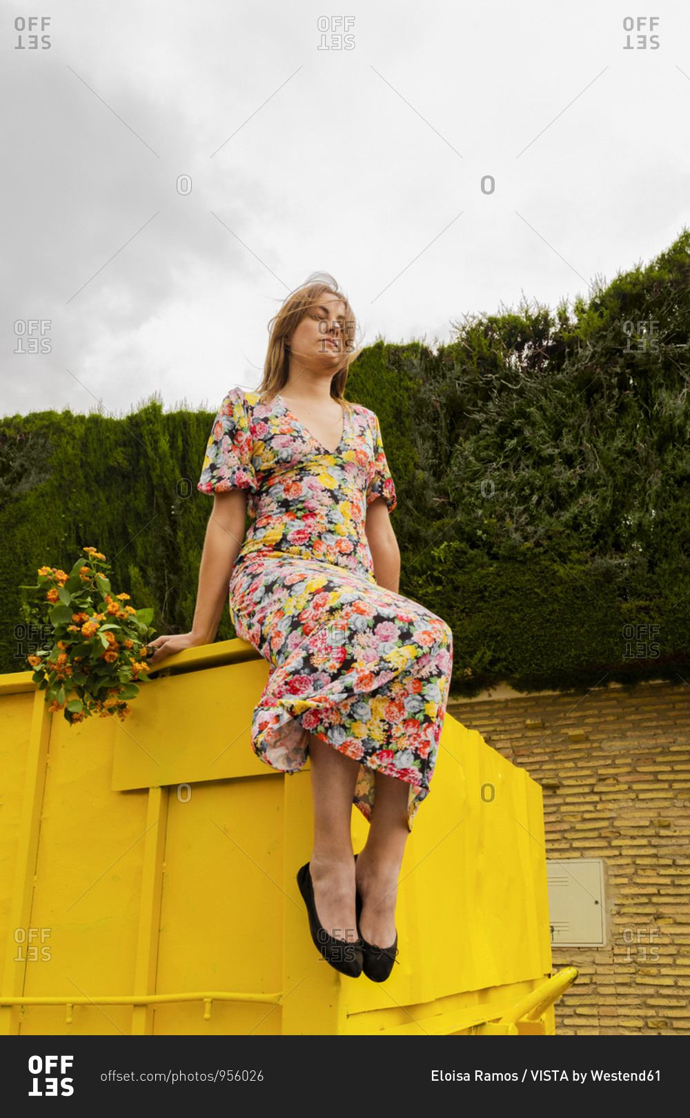 Woman in flower dress sitting on edge of yellow container- holding bunch of flowers