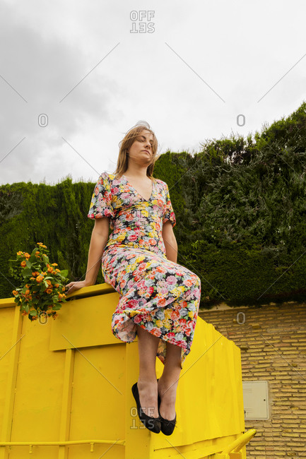 Woman in flower dress sitting on edge of yellow container- holding bunch of flowers