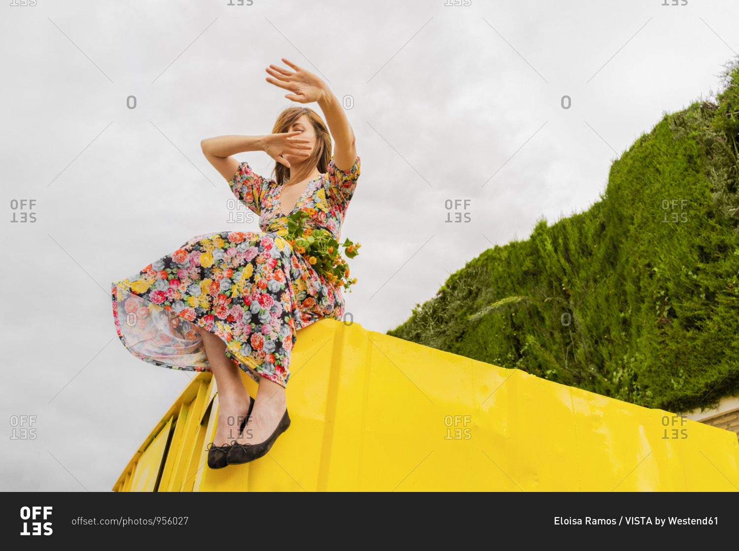 Woman in flower dress sitting on edge of yellow container with bunch of flowers in her lap- moving arms