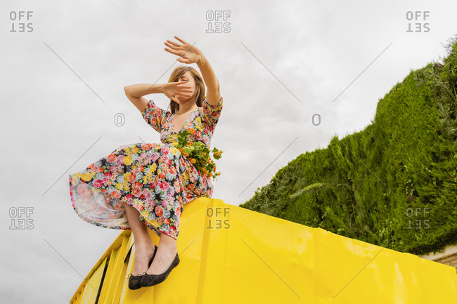 Woman in flower dress sitting on edge of yellow container with bunch of flowers in her lap- moving arms