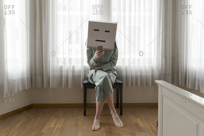 Woman wearing a card box on head with bored smiley sitting on chair and using smartphone