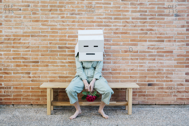 Disappointed woman sitting on bench in front of brick wall with sad face on cardboard box