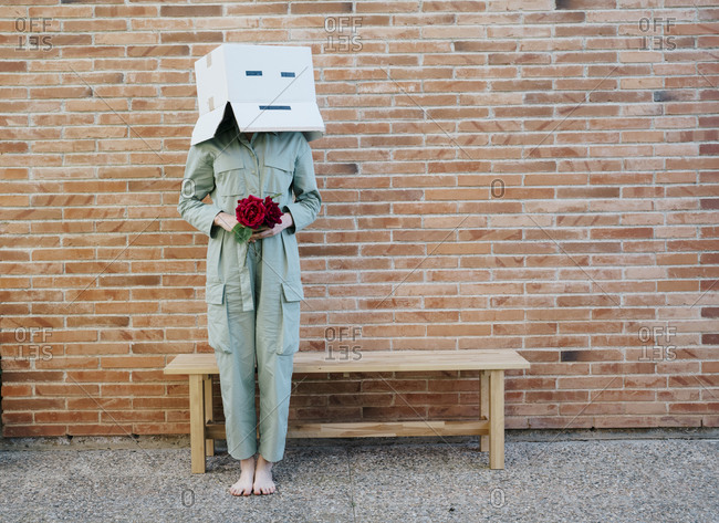 Woman holding red rose- wearing cardboard box with happy face- standing by bench in front of brick wall