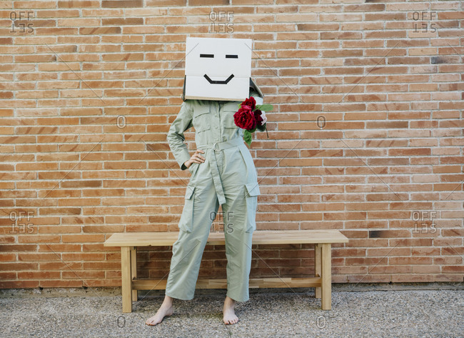 Woman holding red rose- wearing cardboard box with happy face- standing by bench in front of brick wall
