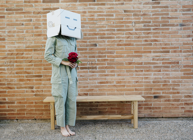 Woman holding red rose- wearing cardboard box with happy face- standing by bench in front of brick wall