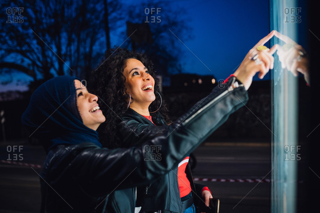 Young woman in hijab with best friend using illuminated touchscreen at city bus stop at night