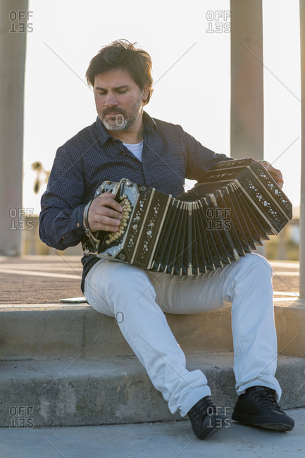 Senior male musician sitting on stone parapet on street and playing accordion while looking away