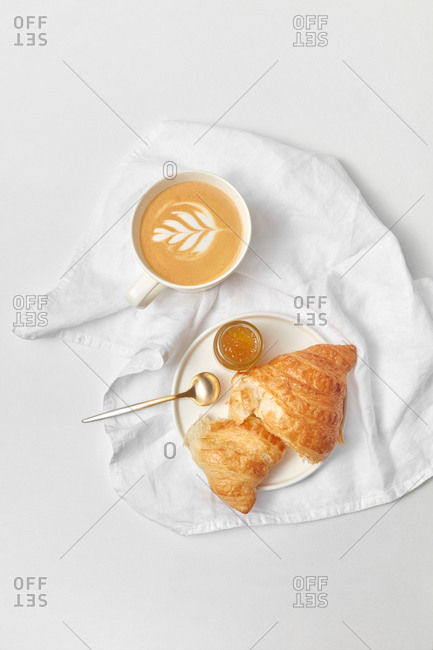 Homemade croissant with jam and cup of cappuccino on a white textile towel and light grey marble bakground, copy space. Top view. Continental breakfast concept.