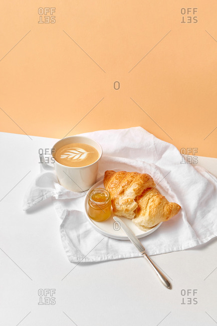 Freshly brewed cappuccino drink and ceramic plate with homemade croissant and jam on a textile white towel on a duotone background, copy space.