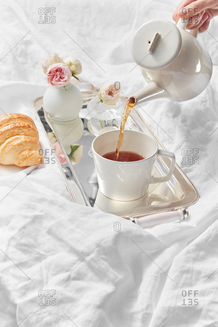 Process of pouring freshly brewed tea in a cup by female's hand for romantic breakfast with homemade croissant and vase with rose on a textile background, copy space.