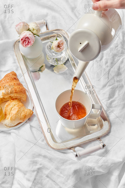 Process of pouring freshly brewed tea in a cup by woman's hand for romantic breakfast with sweet homemade croissant and vase with rose on a silver tray on a textile background, copy space.
