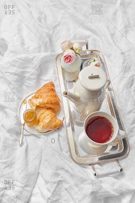 Morning romantic breakfast with tea pot and cup filled hot drink on a silver tray, freshly baked sweet homemade croissant with jam jar on a plate on a crumpled textile sheets, copy space.