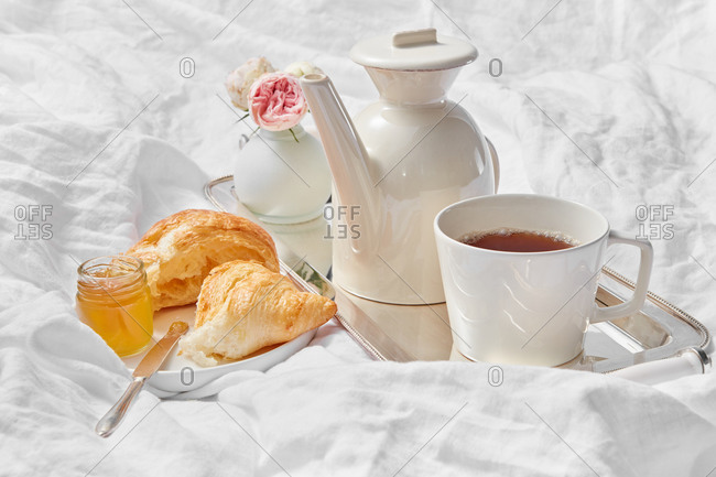 Morning romantic still life with porcelain tea pot and cup filled hot drink on a silver tray, freshly baked homemade sweet croissant with jam jar on a plate on a crumpled textile sheet, copy space.