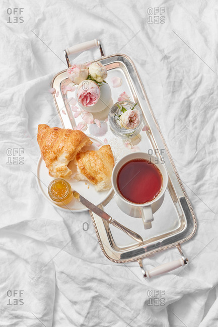 Top view of morning romantic breakfast with ceramic cup filled hot freshly brewed drink on a silver tray and homemade sweet croissant with jam jar on a crumpled textile background, copy space.