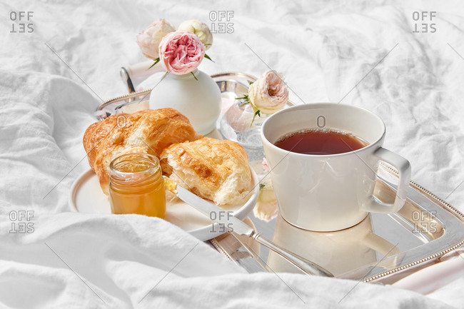 Beautiful romantic still life - silver tray with cup filled freshly brewed hot aromatic tea, flowers vase and croissant with sweet apricot jam on a plate on a crumpled textile background, copy space.