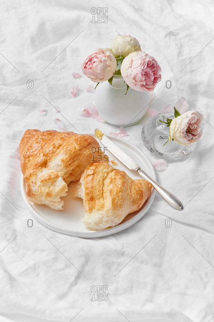 Freshly baked deliciouse homemade croissant on a plate and ceramic vase with fresh pink roses flowers on a textile crumpled background, copy space. Romantic morning breakfast.