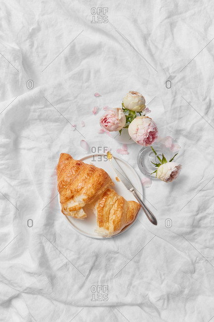 Top view of morning romantic composition with freshly baked homemade tasty sweet croissant on a plate and ceramic vase of aromatic roses flowers on a crumpled textile background, copy space.