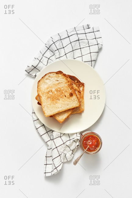 Top view breakfast set with stack of freshly gilled toasts and glass jar of sweet delicious fruits jam on a light grey stone background, served textile towel, copy space.