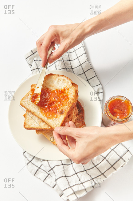 Process of cooking healthy snack or breakfast by female hands from freshly baked toasts and sweet fruits jam on a light grey concrete background, served textile napkin, copy space.