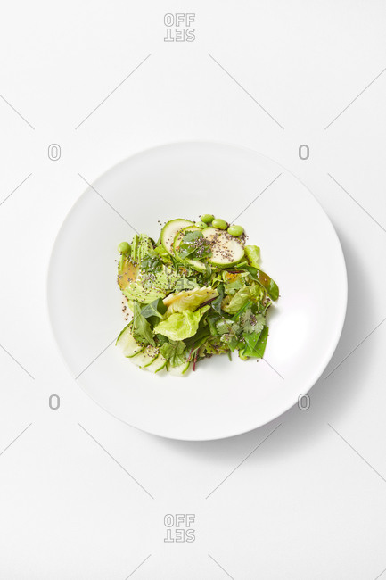 Top view ceramic plate with freshly cooked homemade vegan salad from sliced avocado, zucchini, green leaves and healthy seeds on a white background, copy space. Vegan concept.