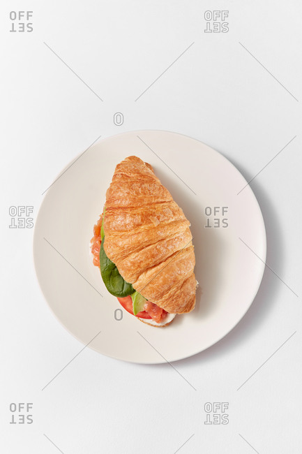 Healthy homemade sandwich croissant with natural salmon fish, green leaf and ripe red tomatoes on a ceramic plate with soft shadows on a white background, copy space.